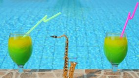 Two bright beautiful yellow-green cocktails with straws glasses, mini alt golden saxophone stand on side of pool, against background of blue water. Summer party, poolside. Slow motion video.