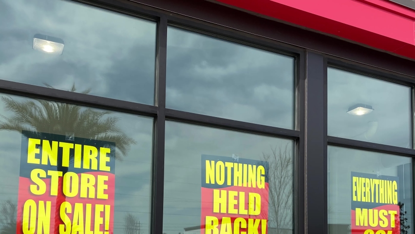 Panning down on signs in the window of a retail store going out of business that say the entire store on sale, everything must go and nothing held back.