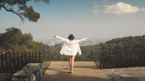 Woman on viewpoint looks on mountain forest landscape, back view. Young female in white goes to cliff observation deck. Summer vacation. Travel destinations, outdoor tourism. Drone flight slow motion