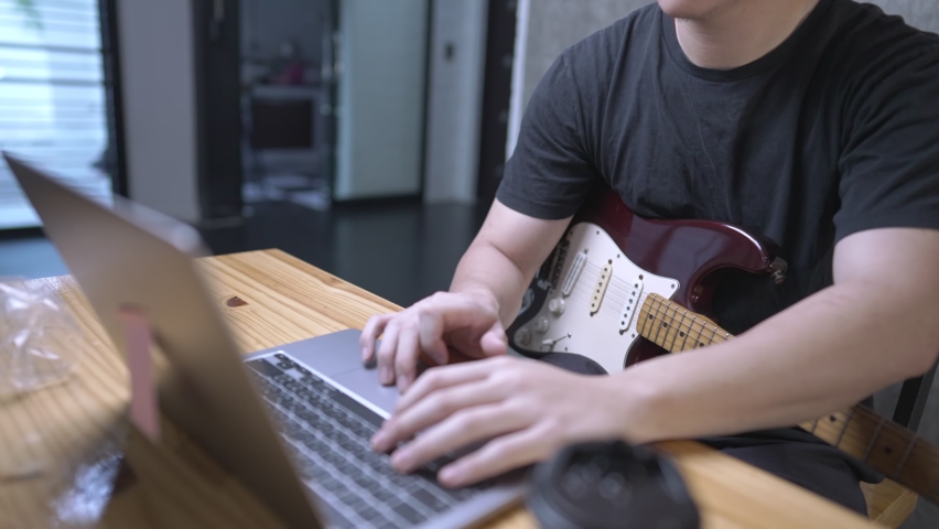 A young white skin man uploading his electric guitar recorded file into portable laptop on wooden office desk, human finger using computer touchpad, freelance guitarist preparing online performance