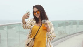Beautiful happy young plus size woman 30-35 years old on vacation takes a selfie on her mobile phone. Smiling overweight female blogger in fashion clothes in yellow color drinks coctail in summer day.