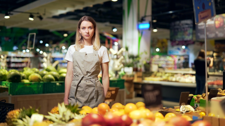 Portrait young woman worker in a Vegetable section supermarket standing smiling with arms crossed. Friendly pleasant female looking at camera in a fruit shop market. Employee in a work apron  Royalty-Free Stock Footage #1077442106