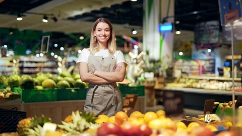 Portrait young woman worker in a Vegetable section supermarket standing smiling with arms crossed. Friendly pleasant female looking at camera in a fruit shop market. Employee in a work apron 