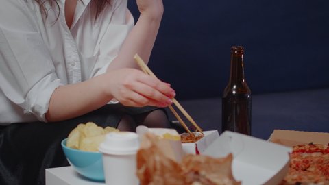 Young woman taking box of chinese food from table to eat using chopsticks while watching movie on television. Person relaxing after work with fast food takeaway in living room at home