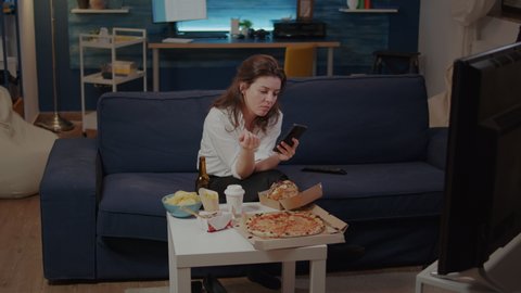 Businesswoman using smartphone while eating slice of pizza from delivery box on table. Young adult browsing on internet, having takeaway food for dinner in living room after work