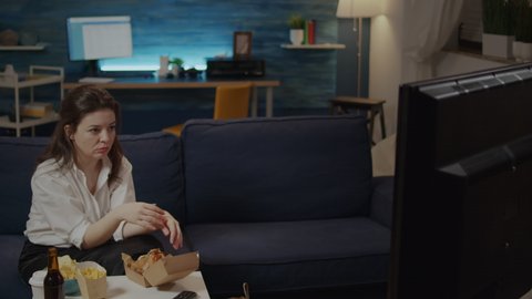 Caucasian woman eating hamburger from delivery bag while watching movie on television at home. Adult enjoying takeaway fast food meal and bottle of beer sitting on couch in living room