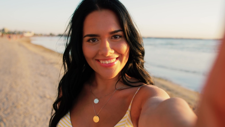 people, summer and swimwear concept - happy smiling young woman in bikini swimsuit taking selfie and blowing kiss on beach