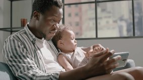 Black father tells story and watches video on smartphone with toddler daughter in grey armchair at big window slow motion