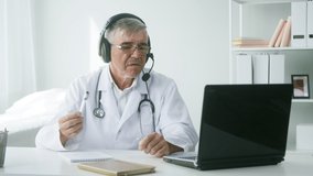 Aged gray-haired doctor in headphones speaking with patient using video call on laptop, male therapist consulting remotely in hospital, elderly physician wearing glasses, medical gown and stethoscope.