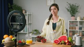 Young woman smiling showing fruits ingredients, food blogger recording video vlog how to cook smoothie made of watermelon, kiwi, citrus and berries. Healthy sweet cocktail. 