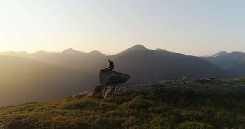 Norwegian with backpack sitting on mountain rock in vivid sunset - drone orbit