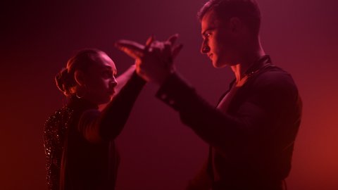 Closeup ballroom couple performing latin dance in red light background. Young dancers looking each other during performance indoors. Beautiful man and woman dancing inside.