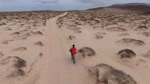 Drone following a young girl on the cycle rides on a white sand road.