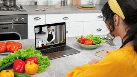 Woman in kitchen study remote culinary lesson listen chef teacher, writes in notebook. Man food blogger in computer screen tells teaches housewife online video call webcam laptop, slices cucumber