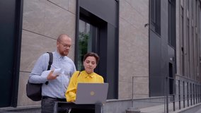 Business people, managers, designers or architectors in casual clothes discussing project presentation outdoors near modern building using laptop and then looking at camera smiling. 4k video footage