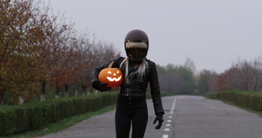 A woman in a black leather suit and a protective biker helmet walks along the road and carries a glowing halloween pumpkin lantern. Halloween is approaching.