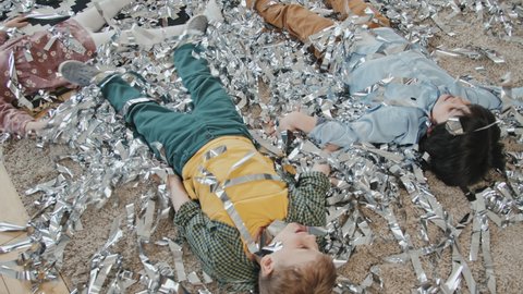 Handheld high angle shot of cute little kids lying on rug littered with silver streamers. They are making snow angles and enjoying themselves