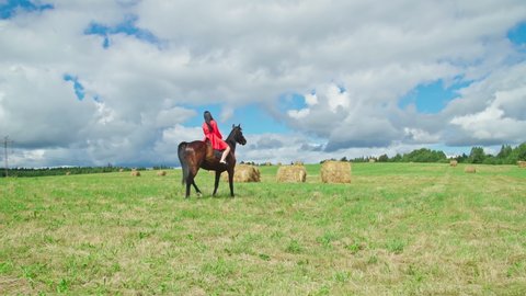 Attractive young woman in red dress riding a horse. Young girl spending outdoor leisure activity, enjoying horseback riding in summer nature. Horseback riding. Back View. 4K