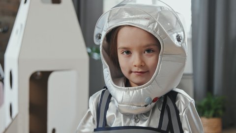 Handheld close up portrait of cute 6-year-old girl in spacesuit putting her visor down and looking at camera. Cardboard rocket ship is in background