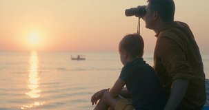 Fun father and son of traveler sitting on sea beach with binoculars under sunset sky in evening time. Family relaxing enjoying holidays, travel day. 4K video