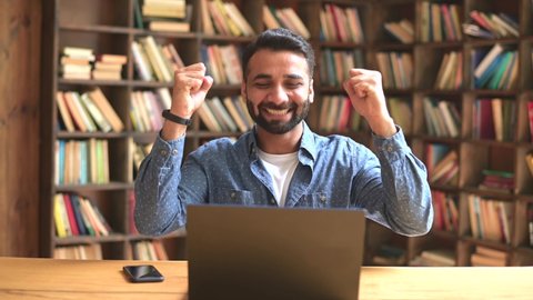Excited surprised middle eastern businessman got unbelievable notification about win, mixed-race indian guy screaming yes, raising fists up in triumph gesture, sits at workplace in loft office