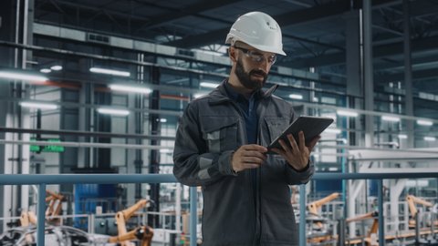 Car Factory Engineer in Work Uniform Using Tablet Computer. Automotive Industrial Manufacturing Facility Working on Vehicle Production with Robotic Arms Technology. Automated Assembly Plant.
