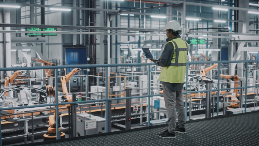 Car Factory Engineer in High Visibility Vest Using Laptop Computer. Automotive Industrial Manufacturing Facility Working on Vehicle Production with Robotic Arms Technology. Automated Assembly Plant. | Shutterstock HD Video #1077496160