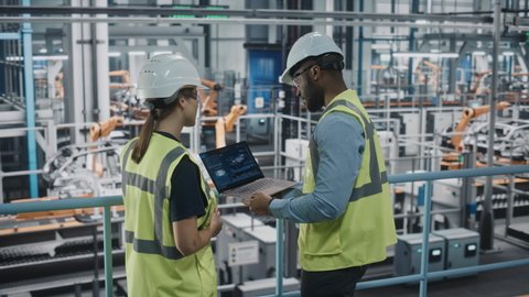 Male Specialist and Female Car Factory Engineer in High Visibility Vests Using Laptop Computer. Automotive Industrial Manufacturing Facility Working on Vehicle Production. Diversity on Assembly Plant.