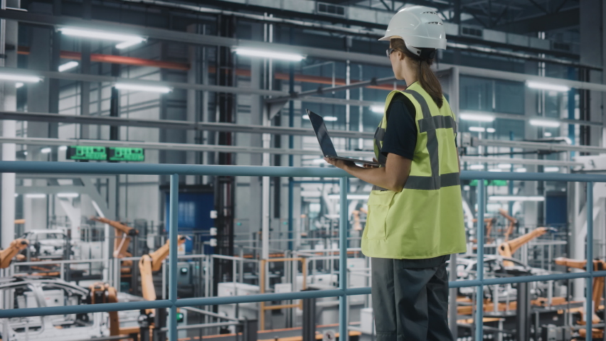 Female Car Factory Engineer in High Visibility Vest Using Laptop Computer. Automotive Industrial Manufacturing Facility Working on Vehicle Production with Robotic Arms. Automated Assembly Plant. Royalty-Free Stock Footage #1077496340