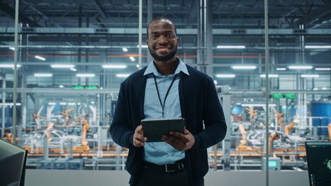 Car Factory Office: Portrait of Successful Black Male Chief Engineer Using Tablet Computer in Automated Robot Arm Assembly Line Manufacturing High-Tech Electric Vehicles. Medium Looking at Camera