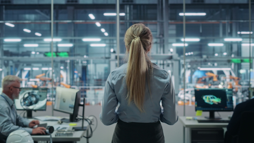 Car Factory Office: Successful Female Chief Engineer Overlooking Factory Production Conveyor. Automated Robot Arm Assembly Line Manufacturing Advanced High-Tech Electric Vehicles. Back View Shot Royalty-Free Stock Footage #1077501893