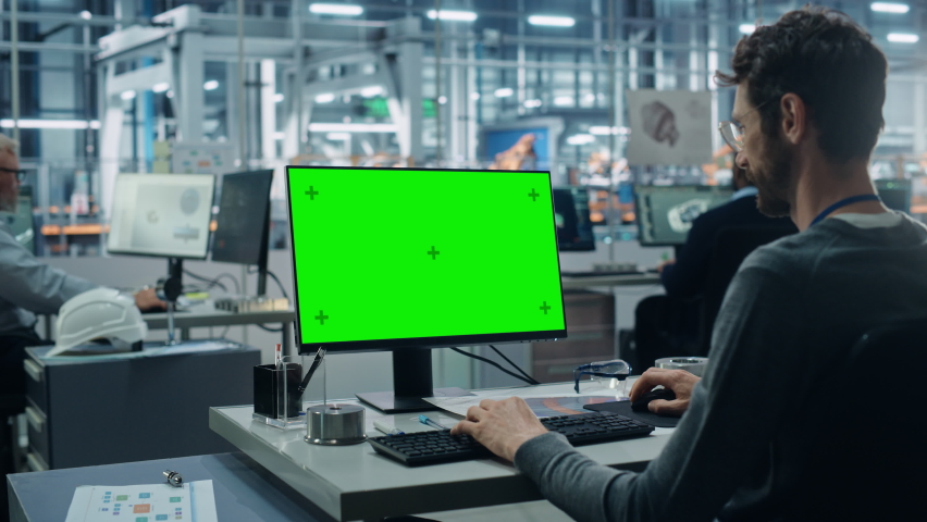 Car Factory Office: Caucasian Male Automotive Engineer Sitting at His Desk Working on Green Screen Chroma Key Computer. Automated Robot Arm Assembly Line Manufacturing. Over Shoulder Medium Shot Royalty-Free Stock Footage #1077501926