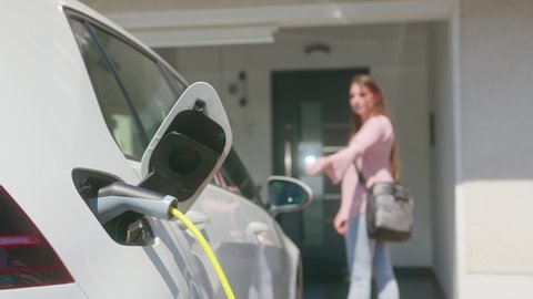 Стоковое видео: Close up of a electric car charger with Caucasian female silhouette in the background, locking a car and entering the home door