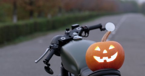 A biker in a helmet picks up a glowing halloween pumpkin lantern from the seat of his motorcycle. Biker and symbols of the holiday of all saints.