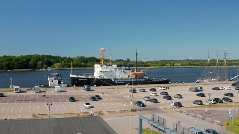 Forwards fly towards icebreaker moored at waterfront. Ship opened to public as museum. Large parking lot nearby. Rostock, Germany in 2021