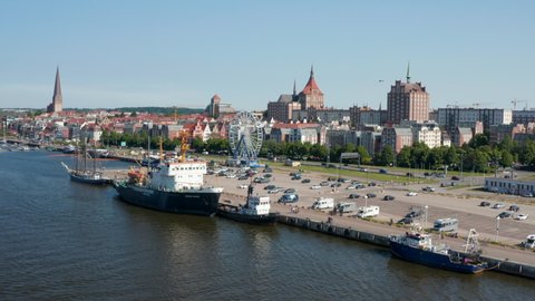 Fly above river along waterfront with parking lot with Ferris wheel amusing attraction. Ships moored at shore, city in background. Rostock, Germany in 2021