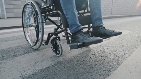 Close-up. Male relative, nursing attendant or medical worker helps non-mobile, paralyzed, non-walking patient to cross road. Modern wheelchair wheel gently slowly rises from carriageway to sidewalk
