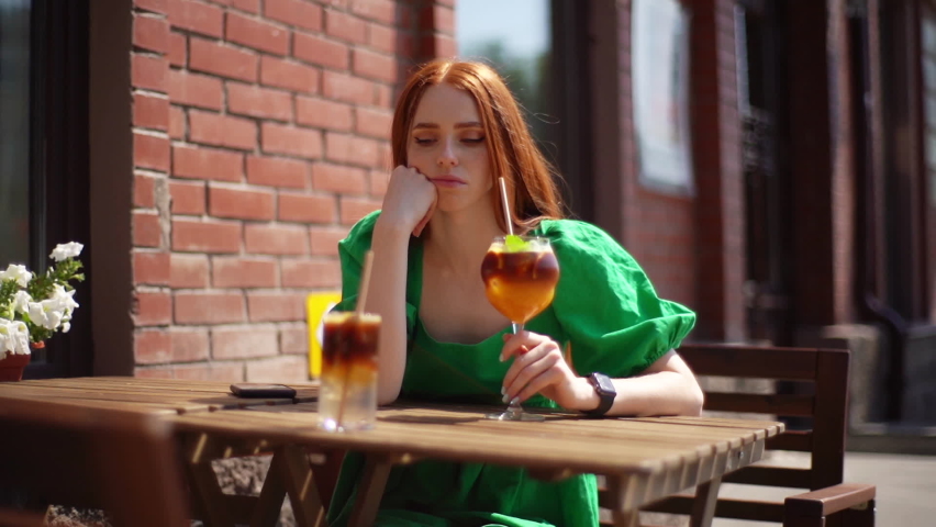 Sad attractive young woman sitting alone waiting for guy on first date, checking time on clock, drinking refreshing cocktail through straw sitting at table in outdoor cafe in sunny summer day.