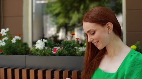 Close-up of happy smiling young woman wearing wireless earphone in ear, using mobile phone sitting at table in outdoor cafe terrace in summer day. Joyful pretty redhead lady listening music.