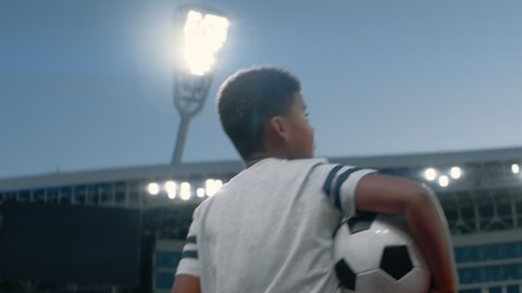 Portrait of African American Black dreamer kid boy walking onto the field of huge soccer football stadium, holding a ball, dreaming of becoming professional player, soccer star