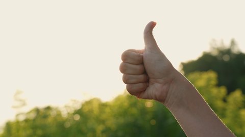 Close-up view 4k video footage of woman giving thumb up gesture cheerfully standing outdoors on sunny sunset time. Female hand with thumb up with sunshine and sunbeams in background