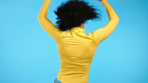 Unrecognizable woman with afro hairstyle dancing positive her back on blue studio background. Pretty female model in yellow wear. Party, happiness, freedom, youth concept.