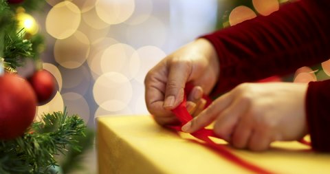 Hands of woman in red dress holding and using ribbon to tie bow for a wrapped gift box for Christmas present with Xmas tree in foreground and colorful lights bokeh in blur background.