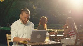A very striking young man is sitting at a table in a public garden, he is doing work on a laptop and looking very happy, in the background two beautiful girls are having a deep conversation