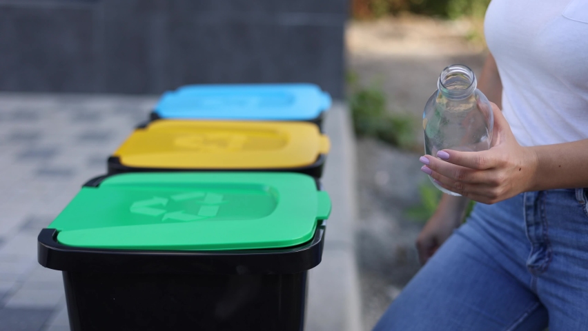 Close-up of woman's hand throwig empty glass bottle in recycling bin. Different colour of plastic bins in the park | Shutterstock HD Video #1077541382