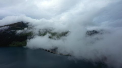 The Roselend dam surrounded by white clouds in Savoie, Auvergne-Rhône-Alpes, France, in the fall, by drone on a day under the clouds.