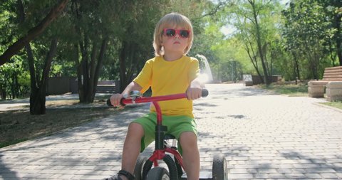 Tranquil little boy kid in stylish glasses rides modern red tricycle along road in green city park on sunny summer day closeup