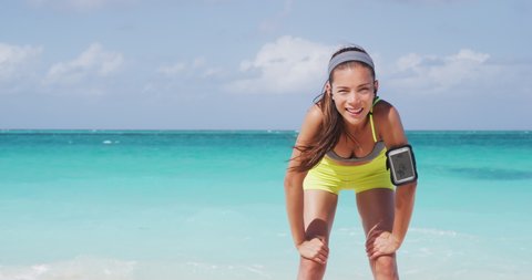 Tired runner listening to music with wireless earphones connected to phone armband holder. Athlete asian woman running on ocean beach outside background