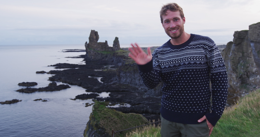 Scandinavian man on Iceland waving hand gesturing hello and welcome to Iceland wearing sweater. Good looking male model looking by ocean sea. SLOW MOTION