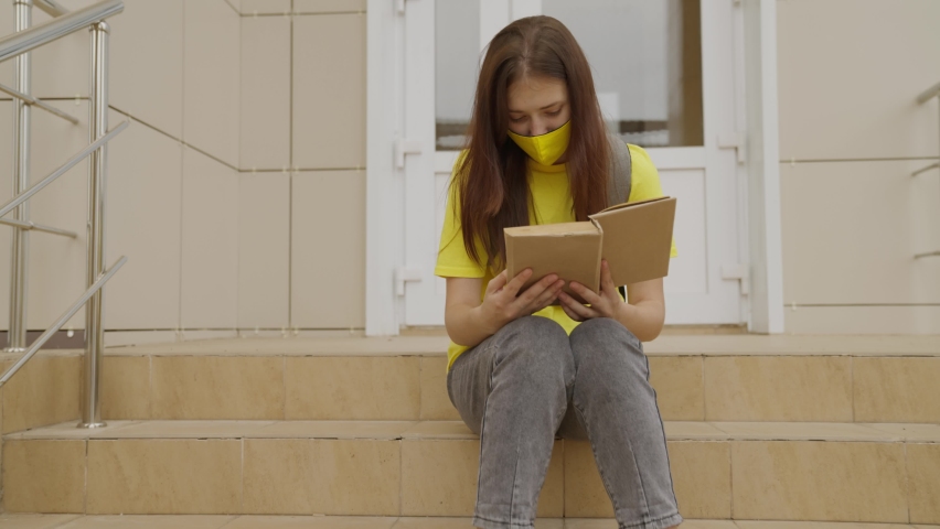 A schoolgirl girl in a mask reads a book while standing at the school, protection against coronavirus, covid-19 pandemic, air filtration through a mask filter, modern education for adolescents Royalty-Free Stock Footage #1077562145
